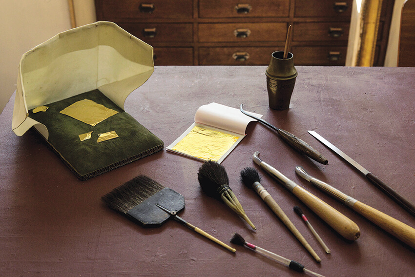Un coussin, une palette à dorer, couteau, pinceaux et brunissoirs sont posés sur une table pour redorer